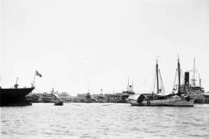 Viaggio in Africa. Porto di Massaua: la poppa di un piroscafo battente bandiera italiana e una imbarcazione a vela ormeggaite a una boa galleggiante. Sullo sfondo le infrastrutture portuali