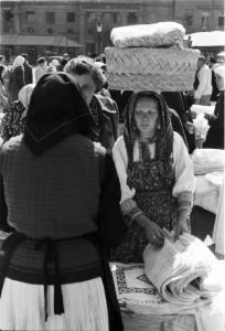 Viaggio in Jugoslavia. Zagabria: mercato. Due donne croate, riprese una da tergo, e una che reca una cesta sulla testa, osservano una bancarella che espone tessuti ricamati