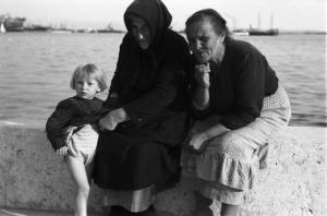 Viaggio in Jugoslavia. Sebenico: coppia di donne anziane con bambina sediti su un muretto nei pressi del porto