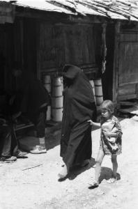 Viaggio in Jugoslavia. Mostar: scene di vita quotidiana - donna con bambina percorre una strada fiancheggiata da baracche in legno