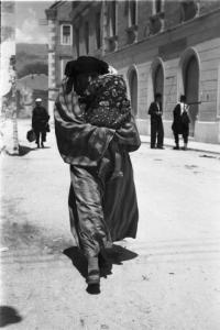 Viaggio in Jugoslavia. Mostar: uomo in abiti tradizionali cammina lungo una strada del paese stringendo tra le braccia un bambino