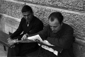 Internamento in Svizzera. Subingen. La famiglia di Don Mangili. Due uomini seduti su una panca appoggiata al muro, uno legge il giornale
