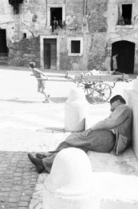 Roma - isola Tiberina. Un giovane ragazzo trascina un carretto al centro di una piazza - in primo piano, all'ombra di un obelisco, dorme un anziano senza tetto