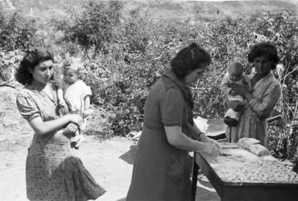 Italia Dopoguerra. Milano - Periferia - Baracche - Donna che prepara gli gnocchi sotto lo sguardo di due giovani madri con in braccio i rispettivi bambini