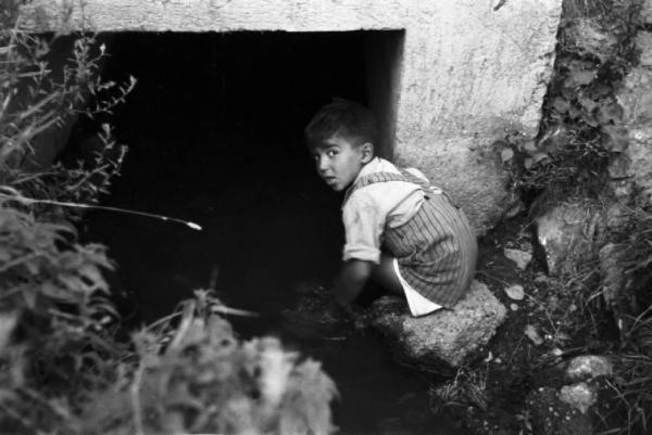 Italia Dopoguerra. Milano - Periferia - Baraccopoli - Ritratto maschile, bambino con i piedi nell'acqua di un canale