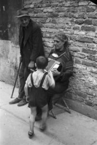 Italia Dopoguerra. Milano - Coppia di anziani mendicanti - la donna suona la fisarmonica mentre un bambino dà un'offerta