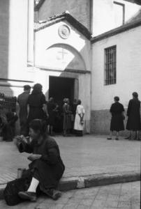 Italia Dopoguerra. Milano - Una donna consuma il proprio pasto su un marciapiede - Alle sue spalle una parrocchia che funge da centro accoglienza