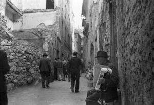 Italia Dopoguerra. Genova - Scorcio di un carrugio con edifici semidistrutti dai bombardamenti - in primo piano un uomo anziano suona una fisarmonica