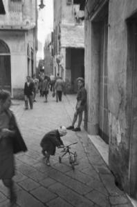 Italia Dopoguerra. Genova - Scorcio di un carrugio - bambino con triciclo tra i passanti