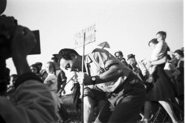 Invasione delle terre. Gruppo di contadini che manifestano. Un uomo pianta un cartello con scritto "la terra a chi la lavora". Si nota la presenza di un cineoperatore