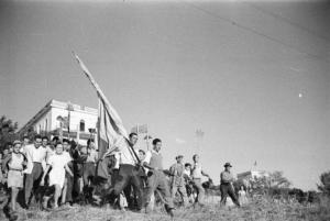 Invasione delle terre. Gruppo di contadini che manifestano marciando in un prato con cartelli, attrezzi agricoli e bandiere