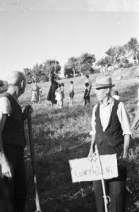 Invasione delle terre. Gruppo di contadini che manifestano in un prato. Due anziani accanto a un cartello con la scritta "W i contadini". Sullo sfondo un prete con bambini intorno