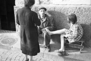 Italia Dopoguerra. Napoli. Un uomo e una bambina seduti lungo il muro di un'abitazione, la bambina appoggia il piede nudo sul ginocchio dell'uomo, mentre una donna parla con lui
