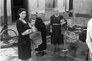 Una donna, una bambina e due anziane attendono il riempimento dei secchi dalle cisterne d'acqua collocate per strada