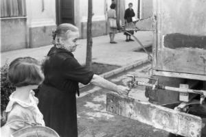 Un'anziana riempie un bicchiere d'acqua da una cisterna collocata per strada - una bambina la osserva