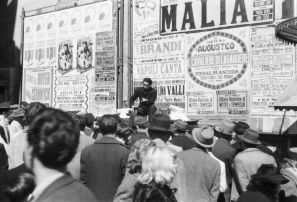 Italia Dopoguerra. Napoli. Folla di persone intorno a un uomo su un calesse in prossimità di cartelloni con spettacoli teatrali e cinematografici