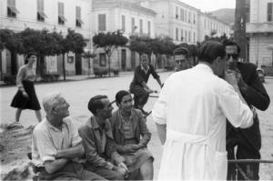 Diano Marina. Dottore in camice bianco parla con la gente del paese in piedi e seduta su una panchina