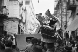 Italia Dopoguerra. Napoli. Folla di gente per le vie della città. Un uomo di spalle trasporta sulla testa un cesto con masserizie tra cui cucchiai in legno