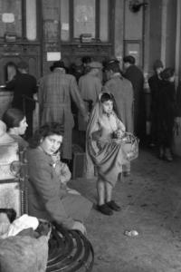 Italia Dopoguerra. Palermo. Madre con bambino seduta su una banchina nei pressi della biglietteria della stazione ferroviaria. Un venditore ambulante bambino. Persone in coda alla biglietteria
