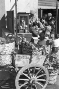 Italia Dopoguerra. Mondello. Bambini intorno al carretto di un venditore ambulante. Carciofi e una piccola teca con statua della Madonna in primo piano