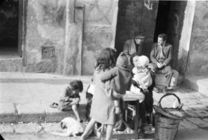 Italia Dopoguerra. Palermo. Bambini attorniano una donna con banchetto lungo la strada. Due uomini discutono seduti sull'uscio di casa