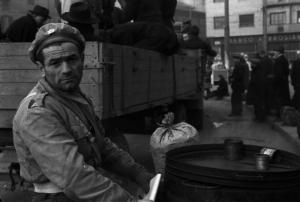 Italia Dopoguerra. Milano. Un venditore di caldarroste ritratto mentre persone si accalcano per salire su un camion