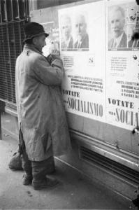 Milano. Un uomo con vestiti strappati si appunta i nomi di due esponenti politici copiandoli dai manifesti elettorali del Partito Socialita affissi ai muri