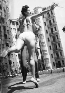 Giovane ragazza in bikini esegue passo di danza seguita dal coreografo. Palazzi sullo sfondo