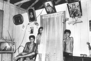 Brasile. Interno di abitazione con due ragazzi e una ragazza - alle pareti sono quadri e santini religiosi e foto di famiglia - a lato un televisore acceso