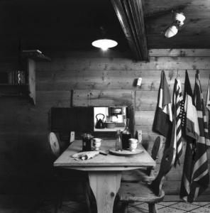 Val Bregaglia. Interno di abitazione rustica con tavolo e pareti in legno e bandiere appese a una parete