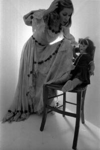 Ritratto femminile - modella in studio  indossa vestito a balze, guarda una bambola seduta su una sedia. Vanessa