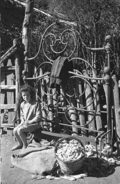 Italia Dopoguerra: Valmontone bombardata. Valmontone - Strada - Ritratto infantile: bambina seduta sulla testata di un letto in ferro