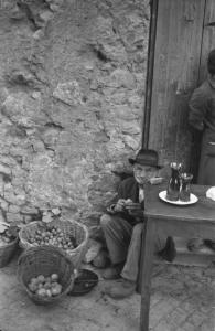 Italia Dopoguerra: Valmontone bombardata. Valmontone - Mercato sulla strada - Ritratto maschile: uomo anziano seduto su un gradino - Ceste di frutta - Tavolo - Brocche
