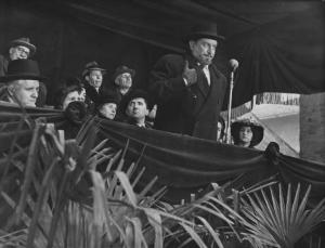 Scena del film "Il cappotto" - Regia Alberto Lattuada - 1952 - L'attore Giulio Stival al microfono su un palco. Dietro di lui un gruppo di attori non identificati
