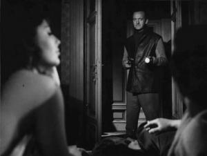 Scena del film "La città prigioniera" - Regia Marco Chiari, Joseph Anthony- 1962 - Un coppia di attori non identificati in primo piano e l'attore David Niven sulla porta con una pistola in mano.