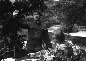 Fotografia del film "Cuore di cane" - Regia Alberto Lattuada 1976 - L'attore Cochi Ponzoni in atteggiamento declamatorio alza un bicchiere presso una tavola imbandita sul bordo di un fiume in compagnia dell'attrice Eleonora Giorgi vista di spalle.
