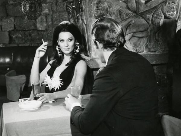 Scena del film "Diamanti che scottano" (L'uomo del colpo perfetto) - Regia Aldo Florio, 1967 - Mezza figura: Rosalba Neri fuma e tiene in mano un bicchiere seduta di fronte a un uomo di spalle.