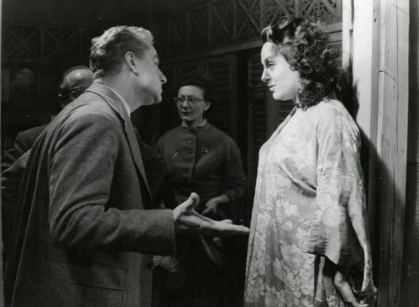 Sul set del film "La diga sul Pacifico" - Regia René Clément, 1957 - Mezza figura di profilo di René Clément mentre istruisce Yvonne Sanson, di profilo, di fronte a lui. Sullo sfondo, un uomo e una donna non identificati.