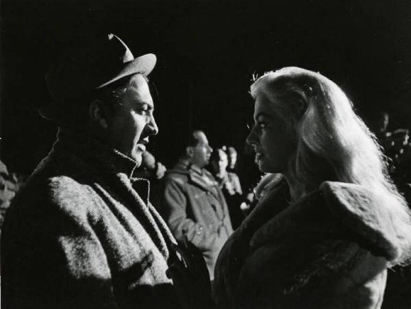 Sul set del film "La dolce vita" - Regia Federico Fellini, 1960 - Mezza figura di profilo di Federico Fellini e Anita Ekberg mentre si guardano. Sullo sfondo, un gruppo di persone.