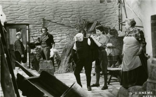 Scena del film "Don Buonaparte" - Regia Flavio Calzavara, 1941 - Totale. Ermete Zacconi viene aiutato da un ragazzo a indossare una giacca. Sullo sfondo Aldo Silvani tiene in una mano un cappello e un bastone e, nell'altra una gallina.