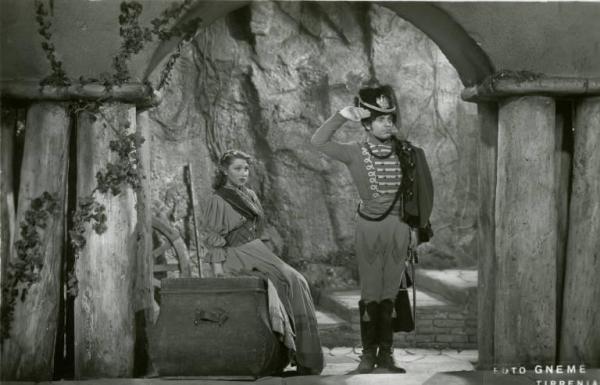 Scena del film "Don Buonaparte" - Regia Flavio Calzavara, 1941 - Totale. Oretta Fiume, seduta su una cassapanca, guarda Mino Doro in piedi accanto a lei, in divisa, mentre fà il saluto militare.