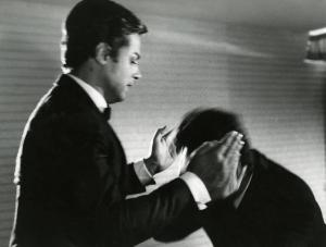 Scena del film "Diabolik" - Regia Mario Bava, 1968 - Mezza figura di John Phillip Law con le mani parallele all'altezza delle orecchie di un uomo, in movimento, chinato dinanzi a lui.
