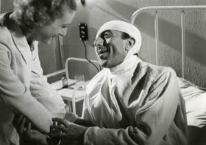 Scena del film "Diagnosi" - Regia Ferruccio Cerio, 1942 - Claudio Ermelli, con la testa fasciata, seduto in un letto di ospedale, porge le mani a un'infermiera che gli sorride.