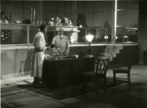 Scena del film "Diagnosi" - Regia Ferruccio Cerio, 1942 - Figura intera di profilo di Franco Scandurra che tiene in mano due quaderni mostrandoli ad un uomo in piedi di fronte a lui.