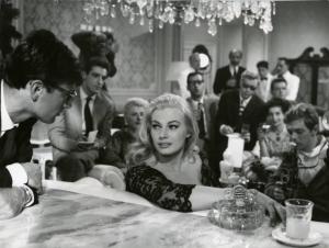 Scena del film "La dolce vita" - Regia Federico Fellini, 1960 - Anita Ekberg, seduta su un divano, appoggia un braccio allo schienale e rivolge il viso verso Sandy von Norman. A destra, Walter Santesso. Attorno giornalisti e paparazzi.