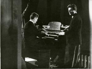 Scena del film "La dolce vita" - Regia Federico Fellini, 1960 - Totale di Alain Cuny, mentre suona l'organo, e Marcello Mastroianni in piedi poggia le mani su di esso.