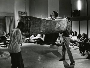 Scena del film "La dolce vita" - Regia Federico Fellini, 1960 - Leo Coleman, a sinistra e Tito Buzzo a destra, sollevano una grande cesta di vimini con dentro Lucia Vasilicò. Un gruppo di persone, sedute in cerchio, li osserva.