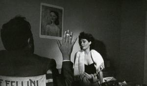 Sul set del film "La dolce vita" - Regia Federico Fellini, 1960 - Federico Fellini, seduto, di spalle, sulla sedia da regista, tiene in mano una sigaretta accesa. Di fronte a lui, è seduta Yvonne Furneaux con un asciugamano sulle spalle.