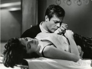 Scena del film "La dolce vita" - Regia Federico Fellini, 1960 - Yvonne Furneaux, sdraiata supina su una barella con gli occhi chiusi. Marcello Mastroianni, seduto accanto a lei, tiene la sua mano appoggiata alla guancia.