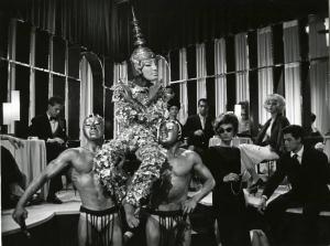Scena del film "La dolce vita" - Regia Federico Fellini, 1960 - Due ballerini, in slip, ne portano un terzo sulle spalle. A destra, Anouk Aimée li osserva. Sullo sfondo, persone sedute ai tavolini, guardano lo spettacolo.